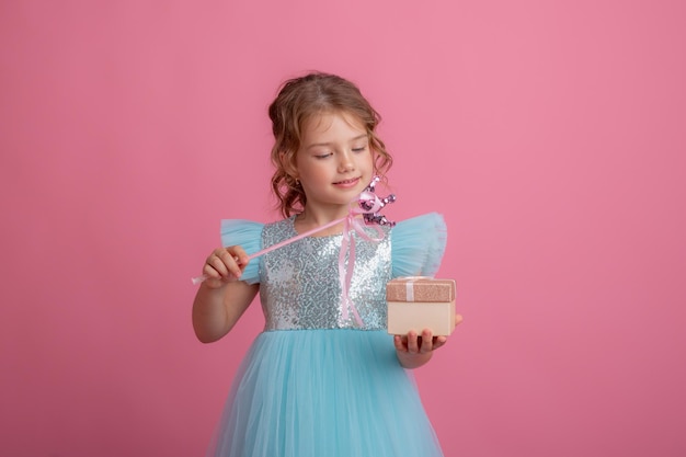 A cute little girl in a beautiful dress holds a fairy's magic wand and a gift on a pink background