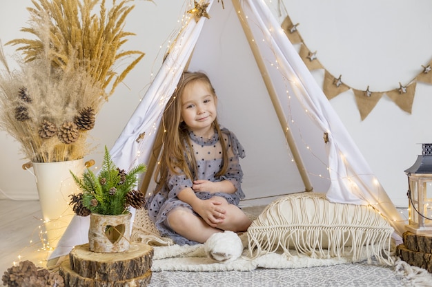 A cute little girl in a beautiful dress plays in a wigwam at home. New Year decoration.