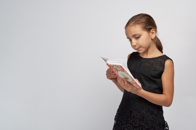 Cute little girl in a black dress with passionate pensive expression reading a book. Large Studio portrait isolated white background