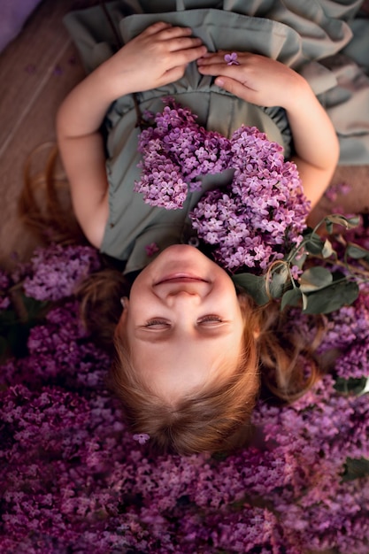 A cute little girl blonde with long hair in a green dress smiles lies with closed eyes among the bunches of lilacs closeup congratulations tenderness memory respect postcard