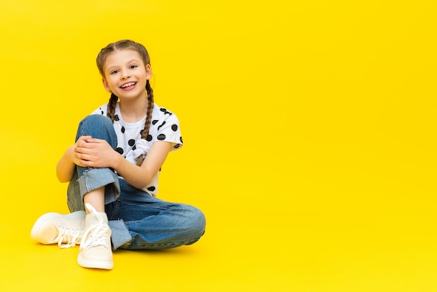 A cute little girl in blue jeans is sitting on a yellow isolated background and smiling