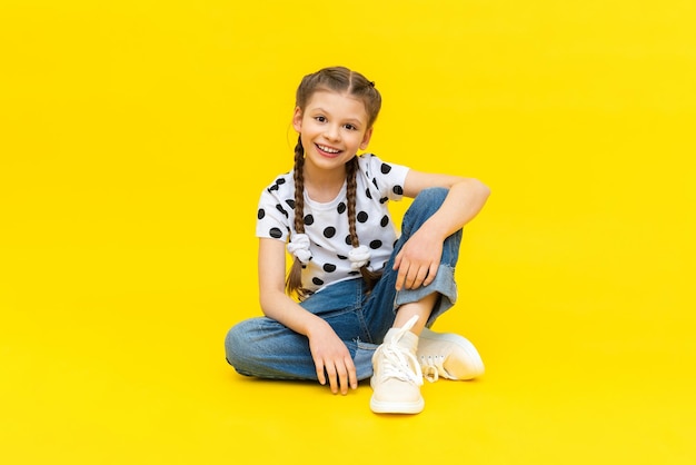 A cute little girl in blue jeans is sitting on a yellow isolated background and smiling