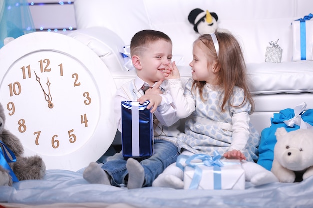 Cute little girl and boy with gifts boxes and big white clock sitting near the white sofa in Christmas and New Year