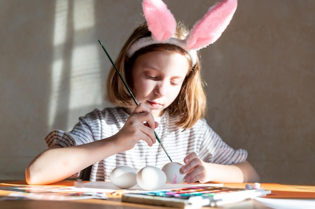 Cute little girl in bunny ears headband painting Easter eggs at table indoorsDIY for easter with kids Craft for spring holidaysdecorations for coloring eggs for holiday Creativity concept