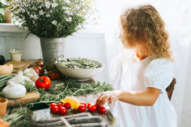 Cute little girl in casual white dress sit near table on veranda of country village house with different vegetables ecofriendly food from garden autumn harvesting