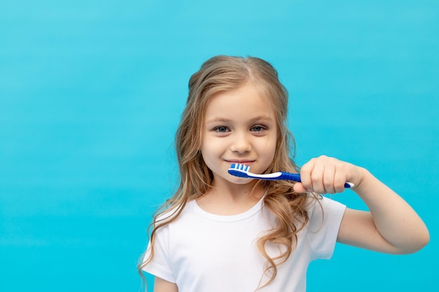 Cute little girl child in a white Tshirt brushing her teeth with a toothbrush on a blue background the concept of hygiene and dentistry space for text