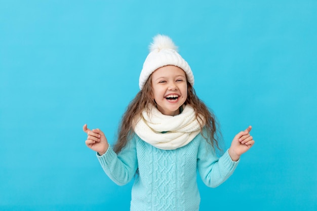 Cute little girl child in winter clothes hat and sweater points fingers up at something on a blue isolated background and smiles and laughs a place or space for text