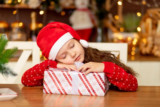 A cute little girl child in winter clothes a Santa Claus hat and a red sweater is waiting for the new year and Christmas having fallen asleep with a gift in the dark kitchen of the house