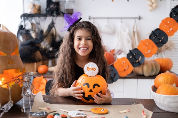 Cute little girl in costume of witch holding pumpkin jack with candies, having fun in kitchen, celebrating Halloween.