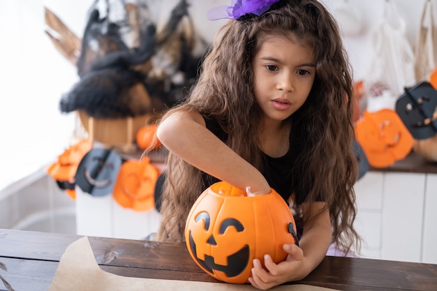 Cute little girl in costume of witch holding pumpkin jack with candies, having fun in kitchen, celebrating Halloween.