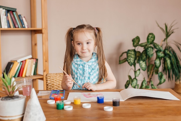 Cute little girl draws with a brush and paints at the table in the living room