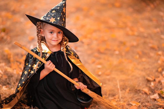 Cute little girl dressed in witch costume celebrating halloween in the park sitting in fallen leaves