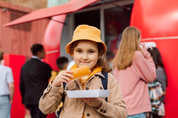 Photo cute little girl eating hot dog nearby food track on a street food delivery or take away food concept