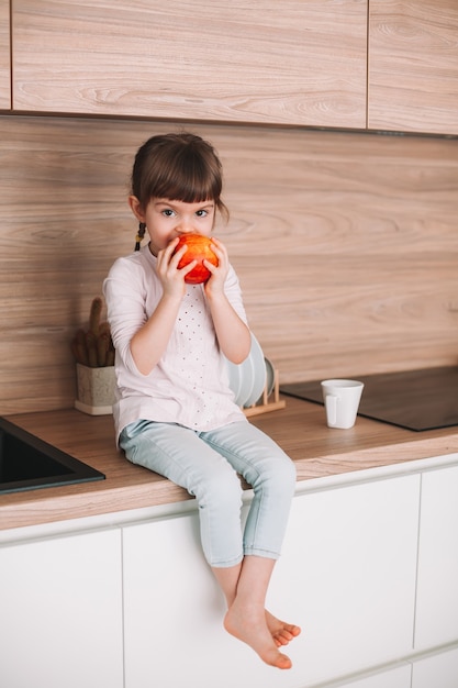 Cute little girl eating juicy red apple sitting on a kitchen surface. Healthy eating concept.