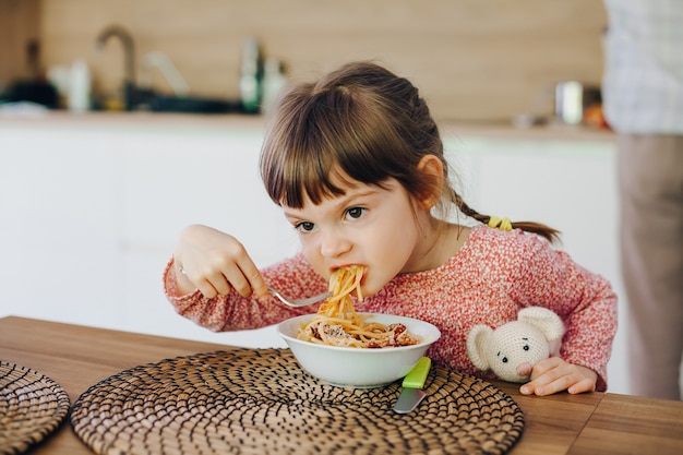 Cute little girl eating spaghetti with sauce sitting in a kitchen at home