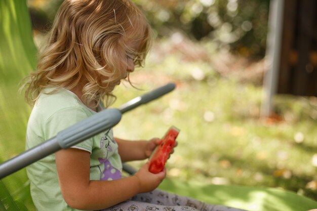 Cute little girl eating watermelon on a deck chair in the garden in summertime