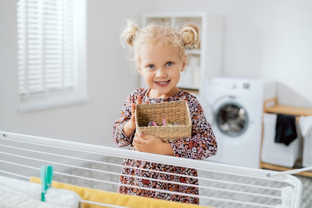 Cute little girl in a flower dress brings a basket of clothes clips stands near the dryer