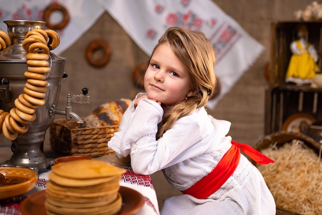 Cute little girl in folk costume seating at the table with samovar while celebrating Maslenitsa