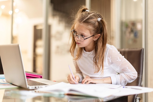 Cute little girl in glasses seriously doing homework using laptop at home for online education, home studying. Kids distance learning. Social distance. Stay at home.