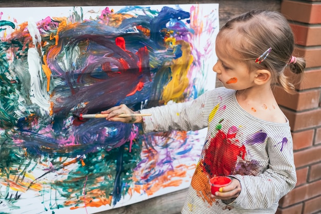 Cute little girl having fun, doing finger painting with various colors on paper on the fence in the yard. Children's creativity art concept.