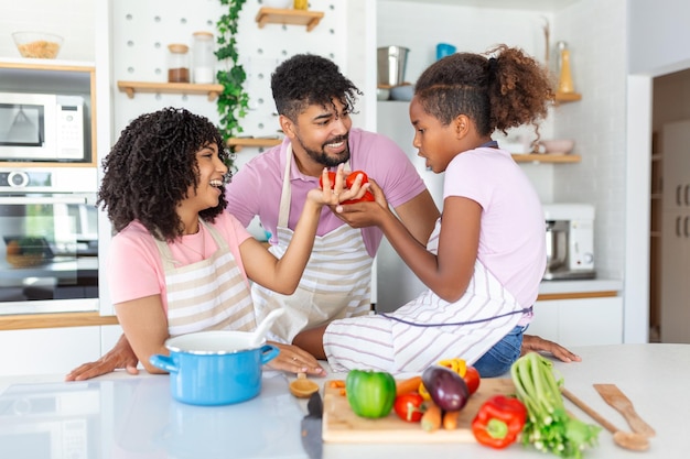 Cute little girl and her beautiful parents are smiling while cooking in kitchen at home Happy african american family preparing healthy food together in kitchen