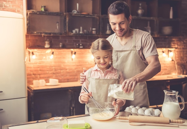 Cute little girl and her handsome dad in aprons are whisking eggs with flour and smiling while baking in kitchen at home