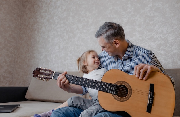 Cute little girl and her handsome father play guitar and smile sitting on the couch at home. father's day. care and education of children.