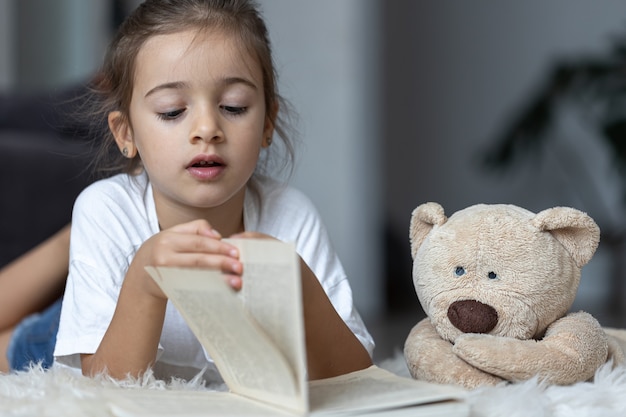 Cute little girl at home, lying on the floor with her favorite toy and reads book.