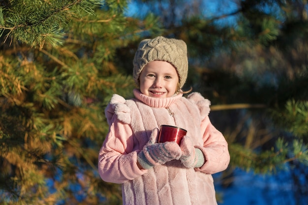 Cute Little girl is Holding a cup with hot tea In Winter forest. Happy childhood. Kids Outdoors.Winter Fun Holiday Concept