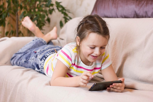 Cute little girl is lying on the sofa at home smiling and using a smartphone