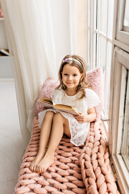 A cute little girl is reading a book on a cozy windowsill.