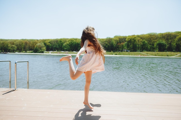 A cute little girl is standing on the pier looking at the beautiful lake child jumping and having fun on the beach