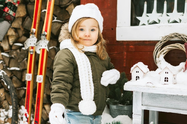 cute little girl in knitted white hat stand at porch of country house and decorate Christmas tree fo