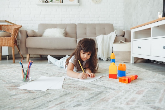 A cute little girl lies on the floor of the house and draws with pencils children's leisure