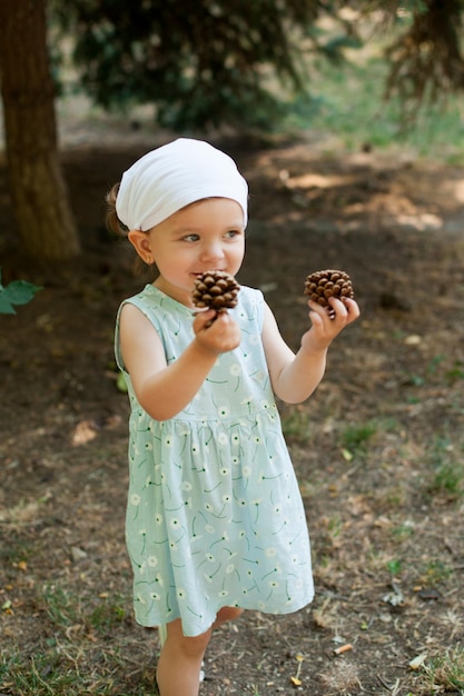 A cute little girl in a light dress walks in the park and collects cones Summer vacation Childhood