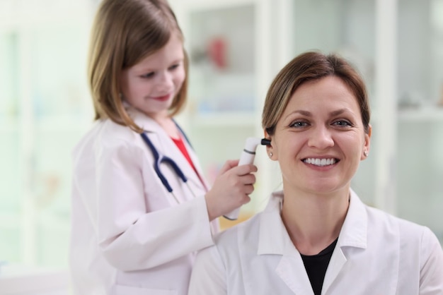 Cute little girl in medical uniform checks ear with otoscope of happy woman curious child