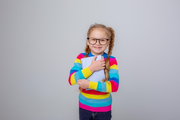 A cute little girl in a multicolored sweater and glasses holds a book on a white background smiling