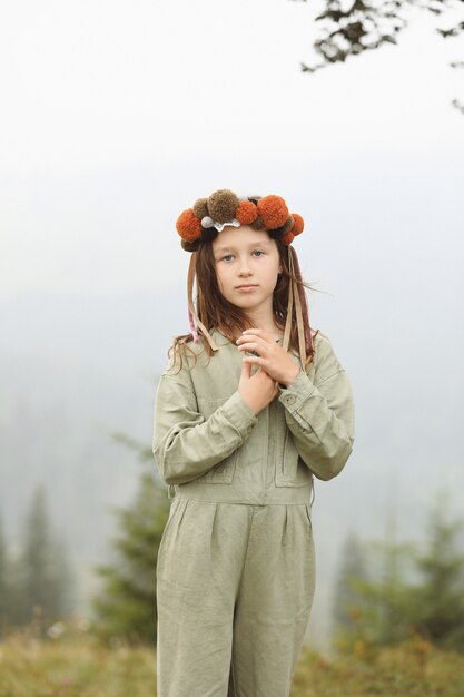 Cute little girl in a pastel jumpsuit with a colorful wreath on her head. Portrait of a little girl outdoors in the foggy mountains