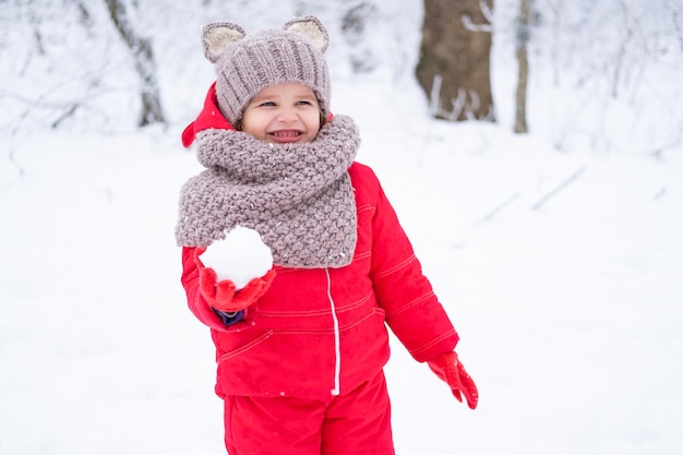 Cute little girl in pink snowsuit and knitted hat and scarf plays with snow in winter forest