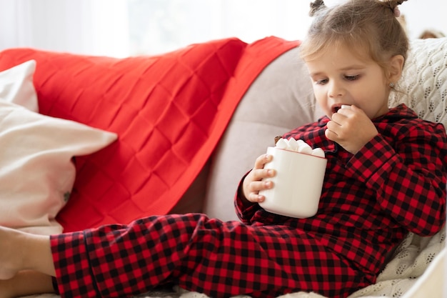 cute little girl in red Christmas pajama holding cup of cacao with marshmallows sitting on couch