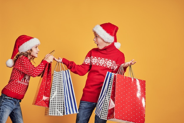 Cute little girl in Santa hat hanging shopping bag with Christmas gifts on hand of astonished boy during holiday celebration against yellow background