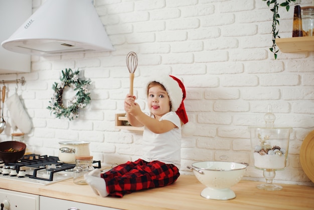 Cute little girl in santa hat, preparing cookies in the kitchen at home. Sits on the kitchen table and helps mom prepare a festive Christmas dinner	