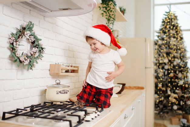 Cute little girl in santa hat, preparing cookies in the kitchen at home. Sits on the kitchen table and helps mom prepare a festive Christmas dinner	