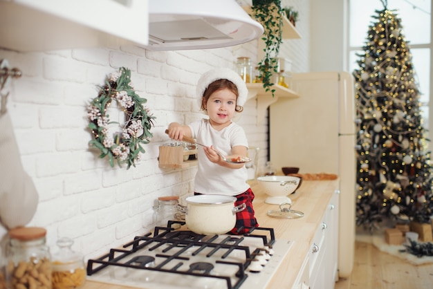 Cute little girl in santa hat, preparing cookies in the kitchen at home. Sits on the kitchen table and helps mom prepare a festive Christmas dinner	