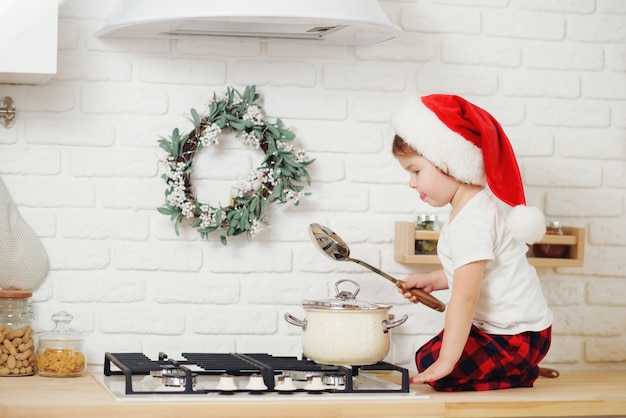 Cute little girl in santa hat, preparing cookies in the kitchen at home. Sits on the kitchen table and helps mom prepare a festive Christmas dinner	