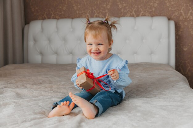 A cute little girl sits on the bed with a gift box at home, accepts birthday greetings.