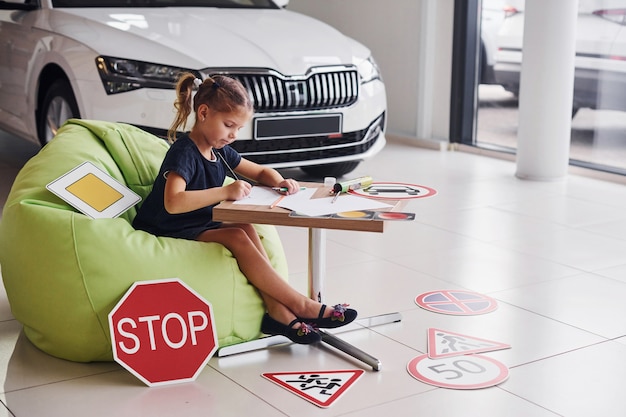 Cute little girl sits on the soft green chair by the table with pencil and paper sheets. Near modern automobile and road signs on the floor.