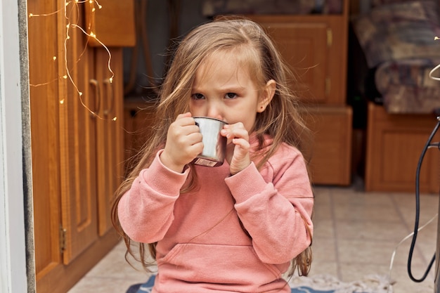 Cute little girl sits on the steps of a travel trailer and drinks hot chocolate with marshmallows