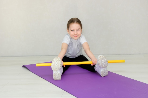 Cute little girl sitting on a mat and doing exercises with a gymnastic stick kids fitness