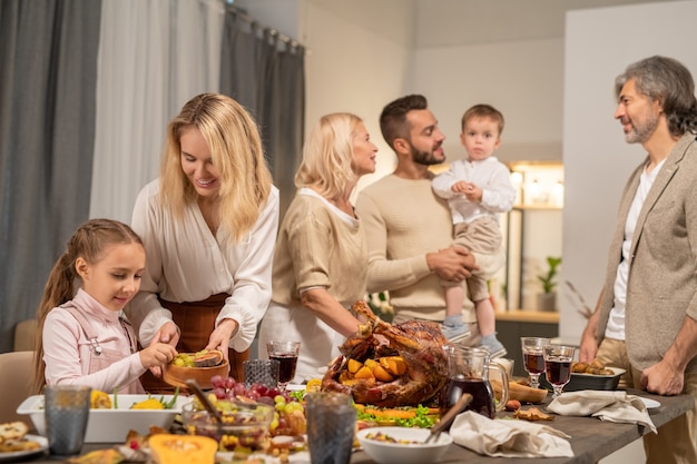 Cute little girl taking fresh grapes from wooden bowl held by her mother on background of grandparents and father with adorable son on hands